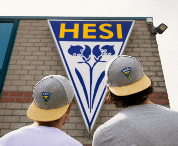 Zwei Personen mit Caps stehen vor einem HESI Logo an einer Wand, ideal für Growshop-Marketing.