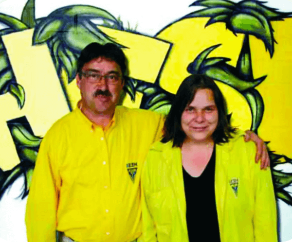 Paar in gelben Hemden vor einer bunten Wand mit Pflanzenmuster, Darstellung von Growshop-Themen.