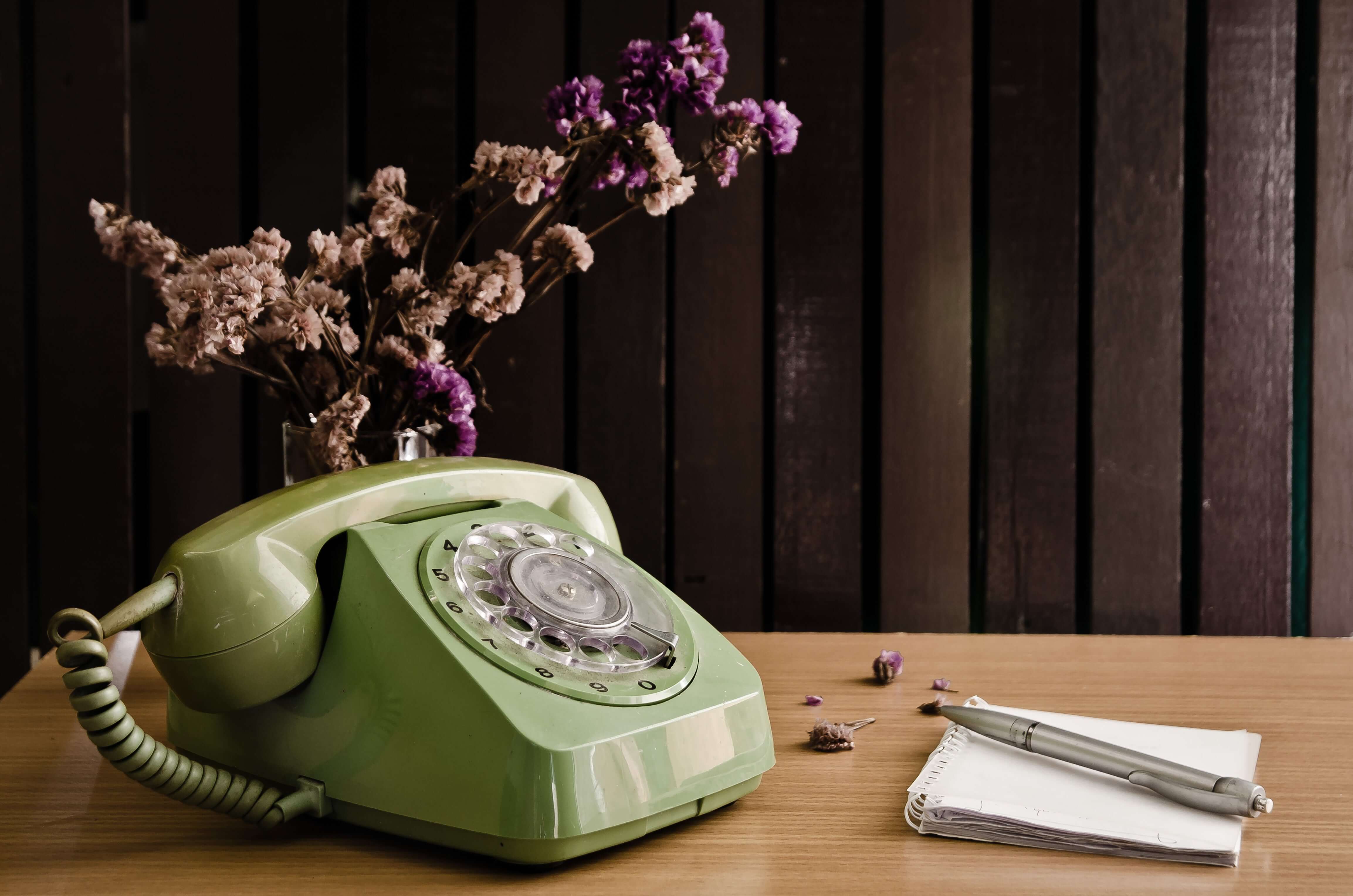 Vintage grüner Telefon, getoppt mit Trockenblumen, auf einem Holztisch neben Notizblock und Stift.