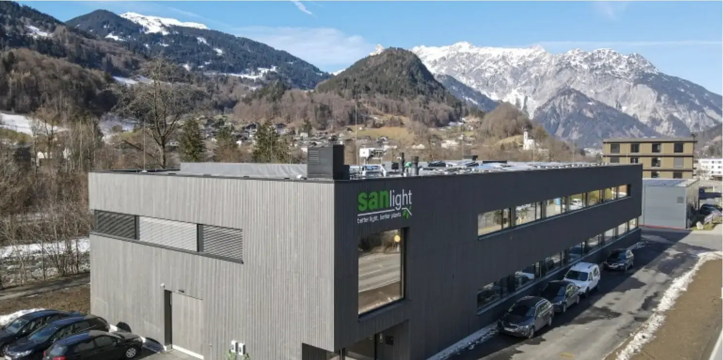 Moderne Firmengebäude mit sanlight Logo, umgeben von Bergen und Autos, in einer schönen Landschaft.